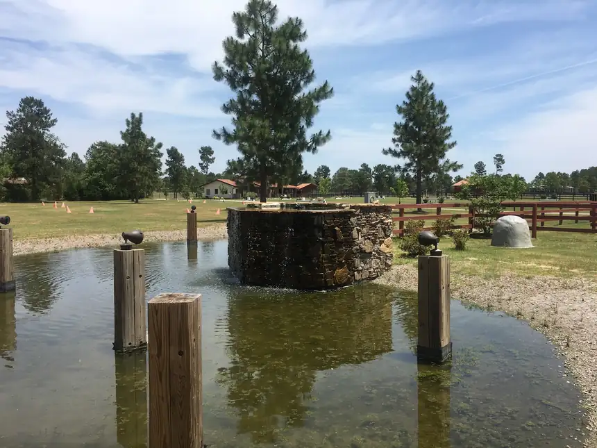 Stone fountain structure standing in a pond with timber posts and pasture behind